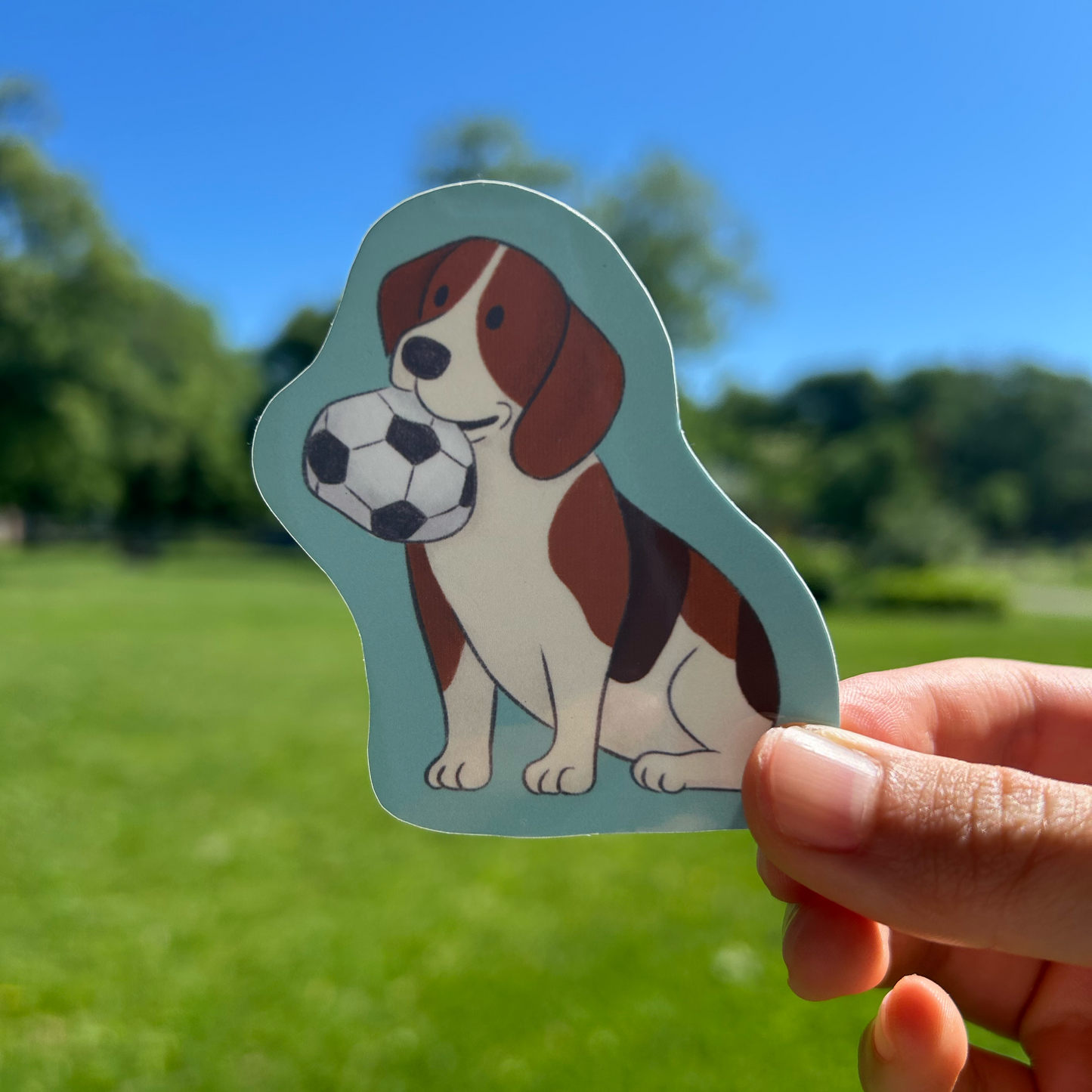 Hand holding a dog-shaped sticker with a soccer ball on a grassy background