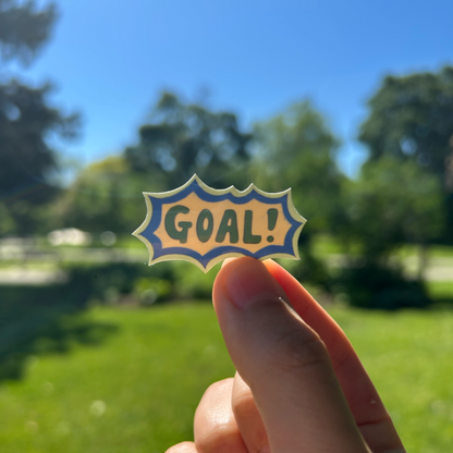 Hand holding a 'GOAL!' sticker outdoors with trees and grass in the background