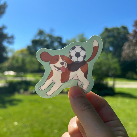 Hand holding a sticker of a dog playing with a soccer ball against a grassy outdoor background