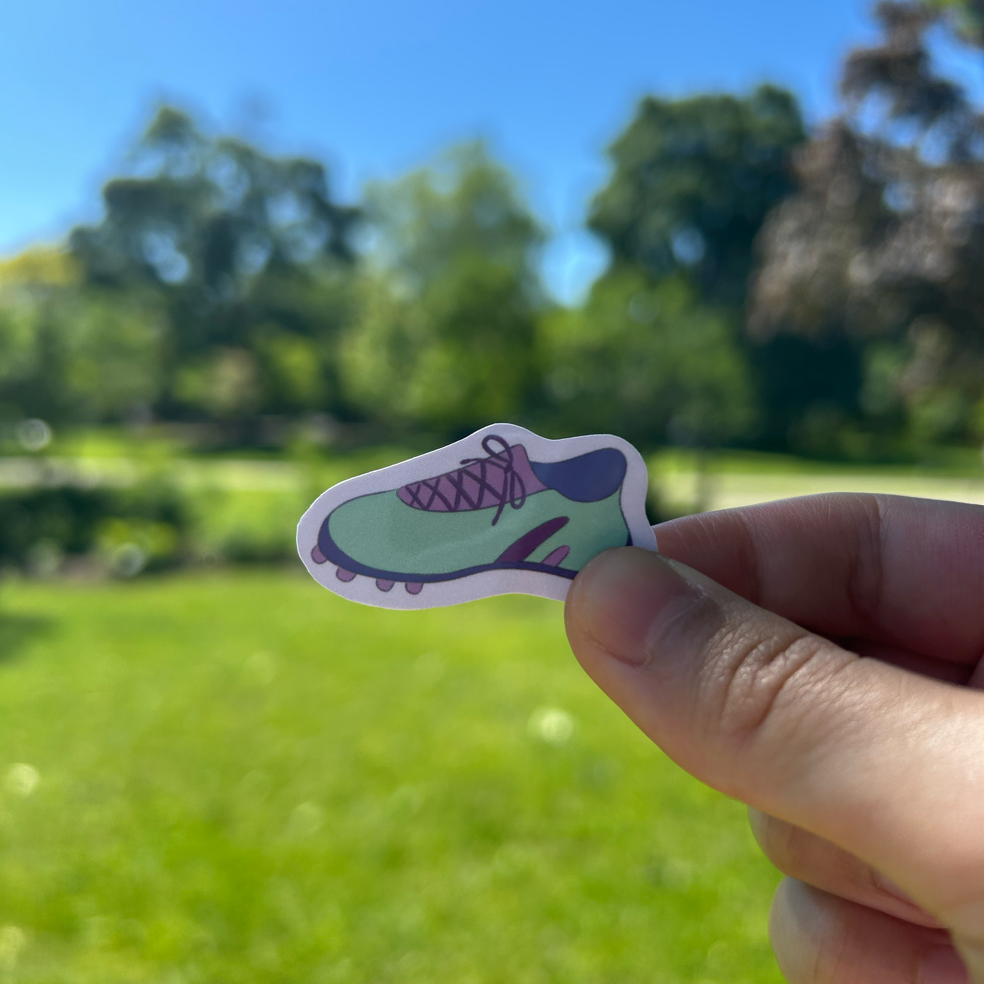 Hand holding a small sticker of colorful soccer cleats against a blurred outdoor background.