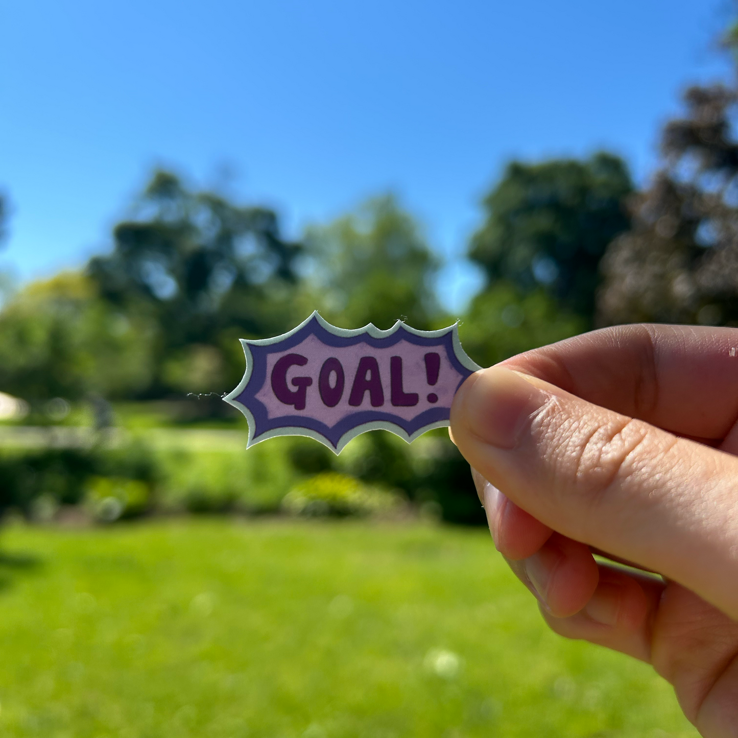 Hand holding a 'GOAL!' sticker against a blurred outdoor background