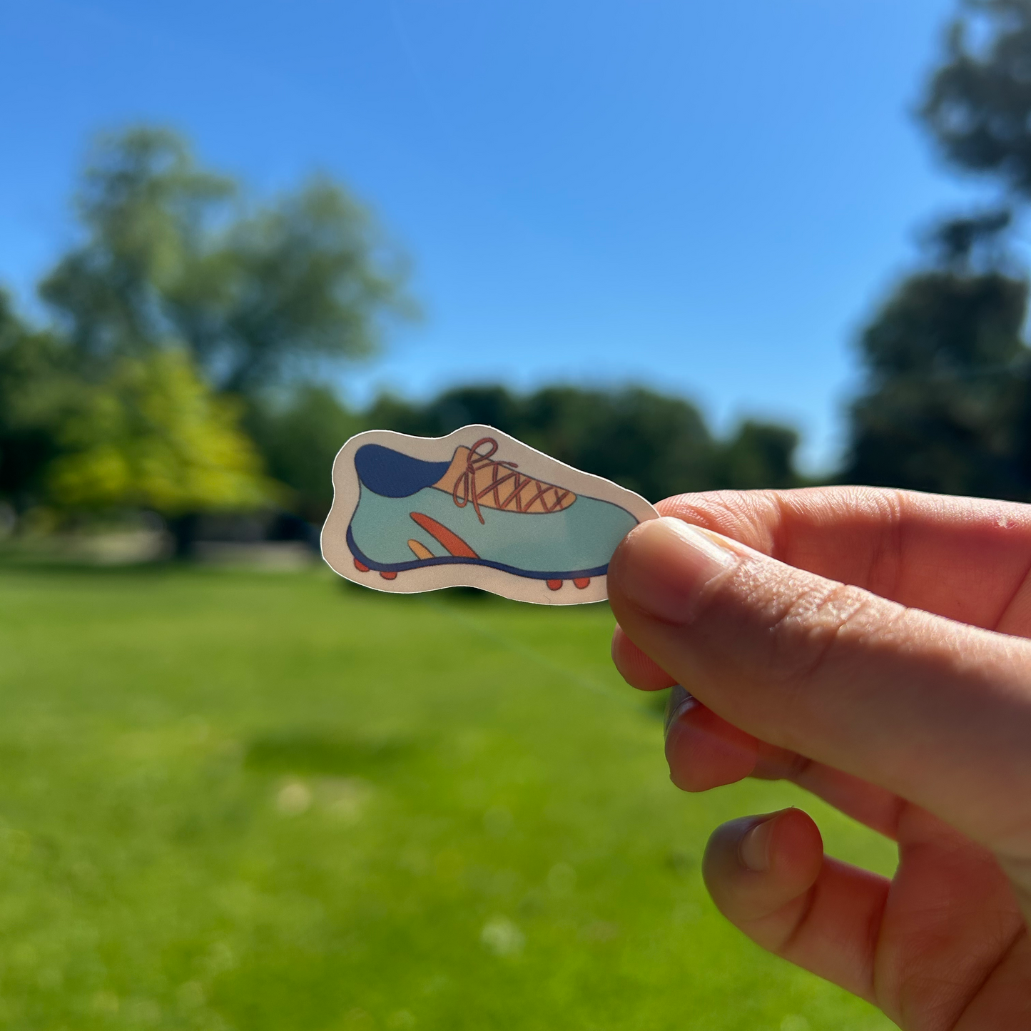 Hand holding a colorful soccer cleat sticker with a blue sky and green grass background
