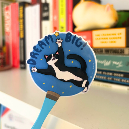 Handheld blue circle sticker with a black and white cat illustration and 'Dream Big!' text, set against a bookshelf background.