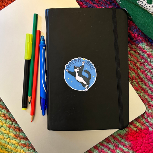 Black notebook with a cat illustration on a colorful textured surface with pens and markers.