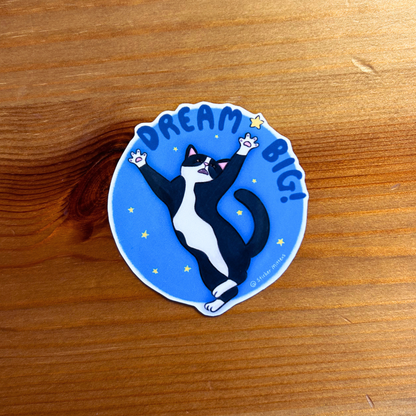 Sticker with a cat illustration and 'Dream Big!' text on a wooden surface