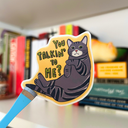 Yellow sticker with a cat illustration and text 'You Talkin' to Me?' against a blurred bookshelf background.