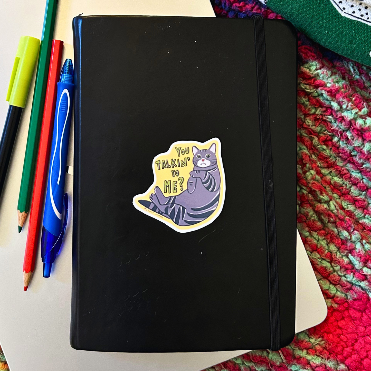 Black notebook with a cat sticker on a colorful knitted surface with pens and pencils.