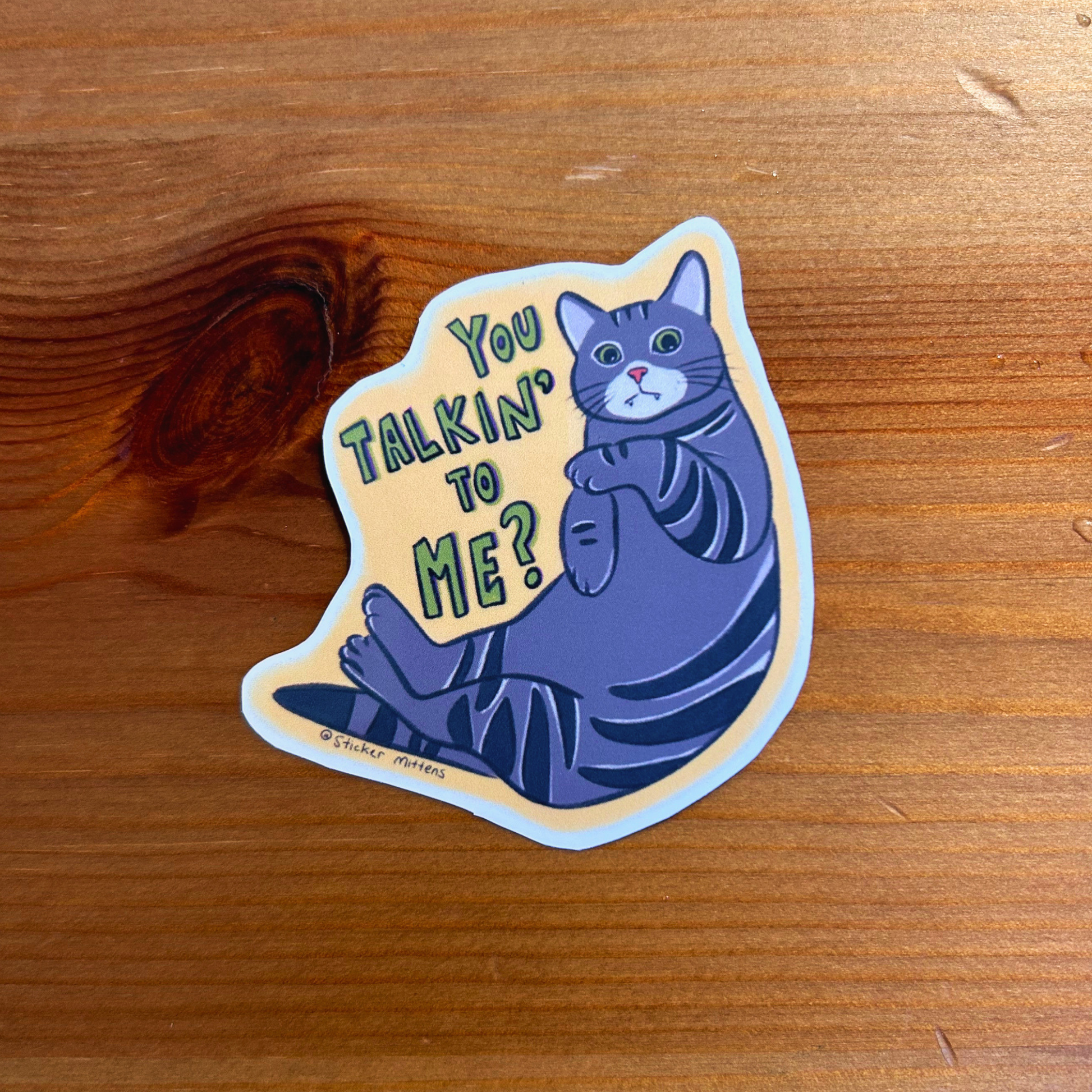 Sticker with a cat illustration and text 'You Talkin' To Me?' on a wooden surface