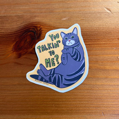Sticker with a cat illustration and text 'You Talkin' To Me?' on a wooden surface