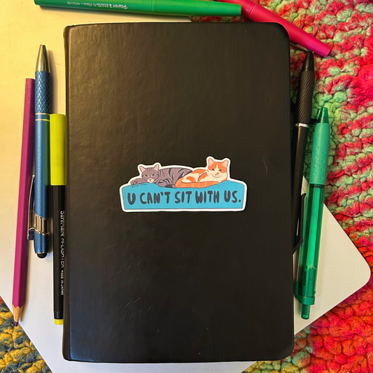 Black notebook with a sticker of two cats and text 'U CAN'T SIT WITH US' on a colorful surface with pens.