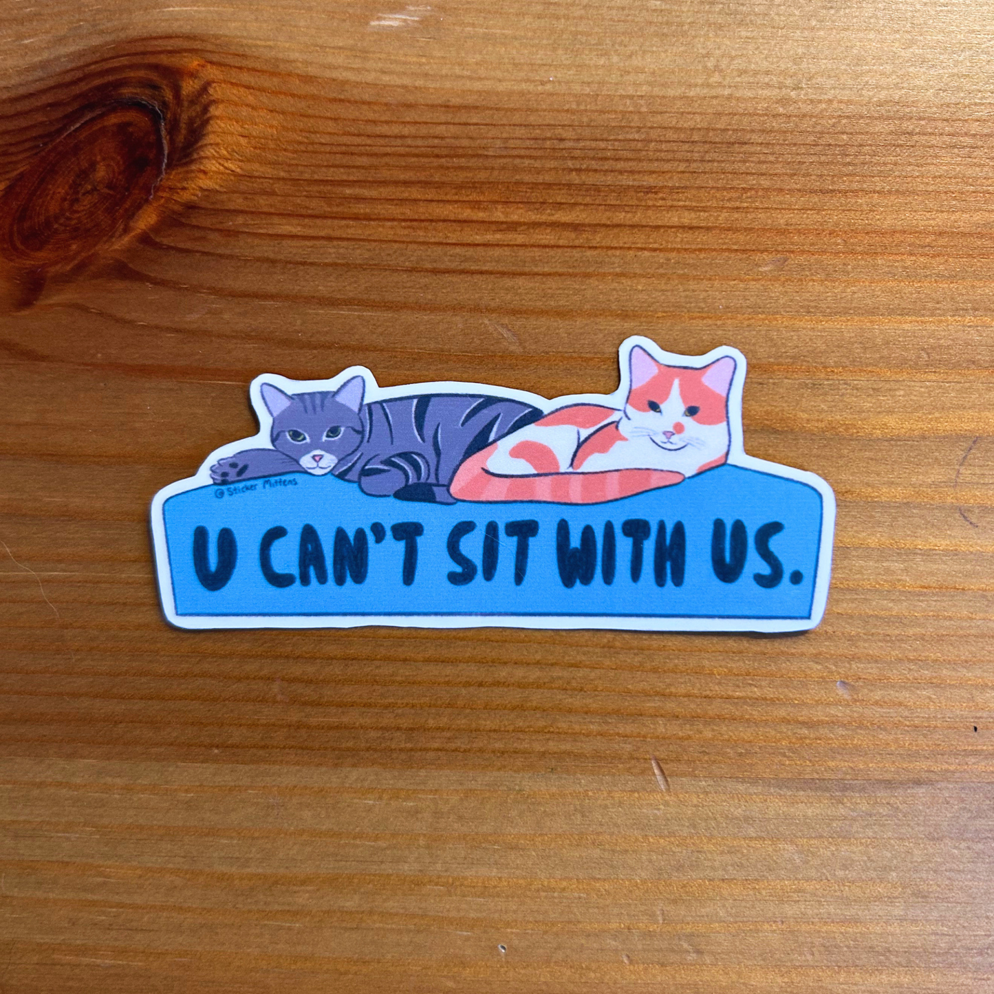Sticker with two cats on a blue bed and text 'U CAN'T SIT WITH US.' on a wooden surface