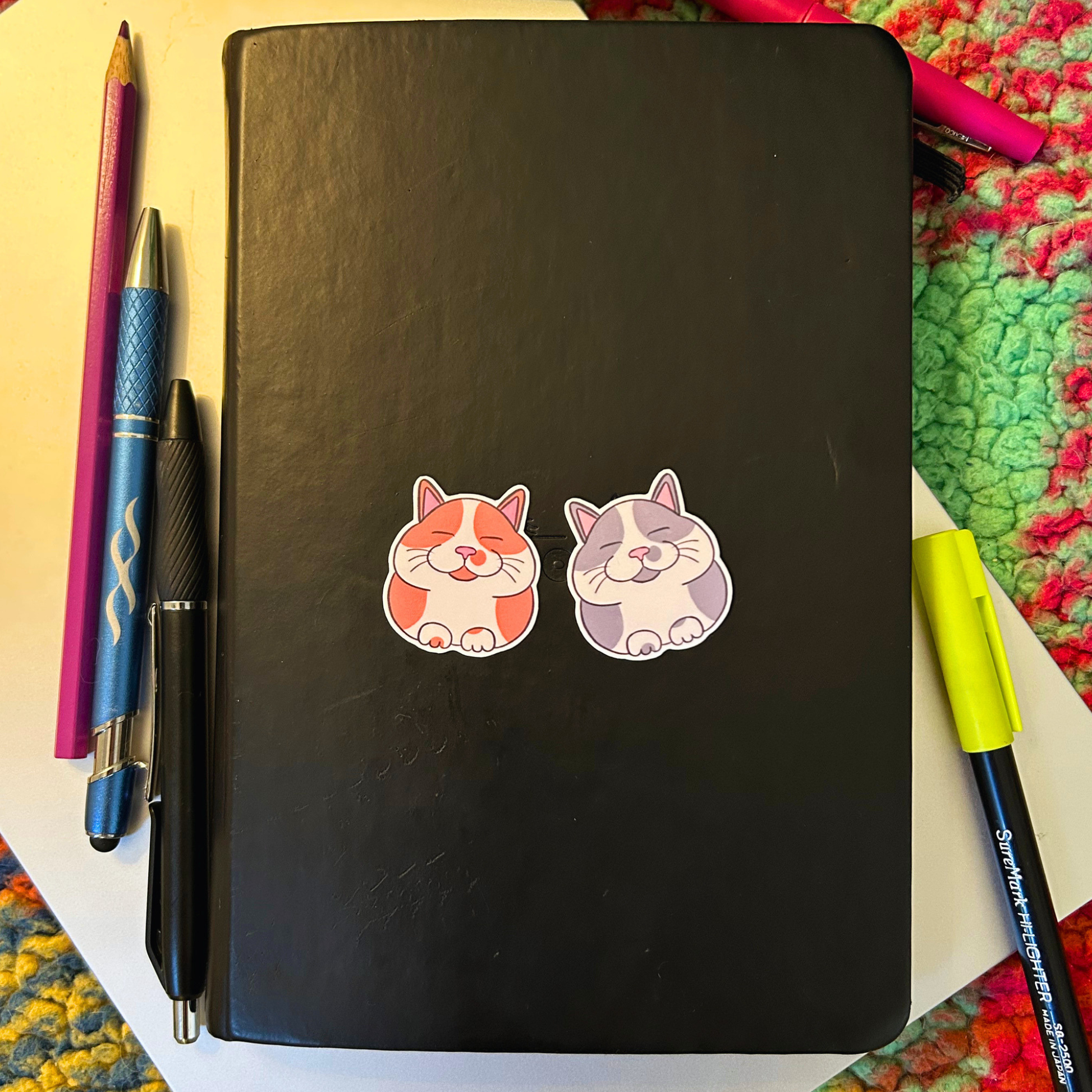 Black notebook with two cat stickers on a colorful surface with pens and pencils.