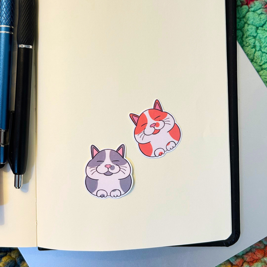 Two cat-shaped stickers on a notebook with pens beside it.