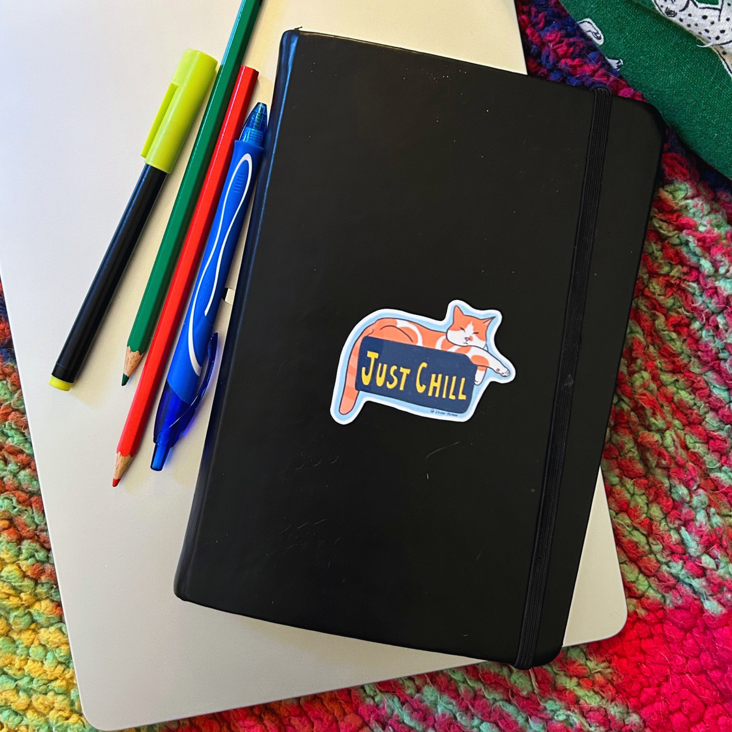 Black notebook with 'Just Chill' sticker on a colorful textured surface with pens and pencils.